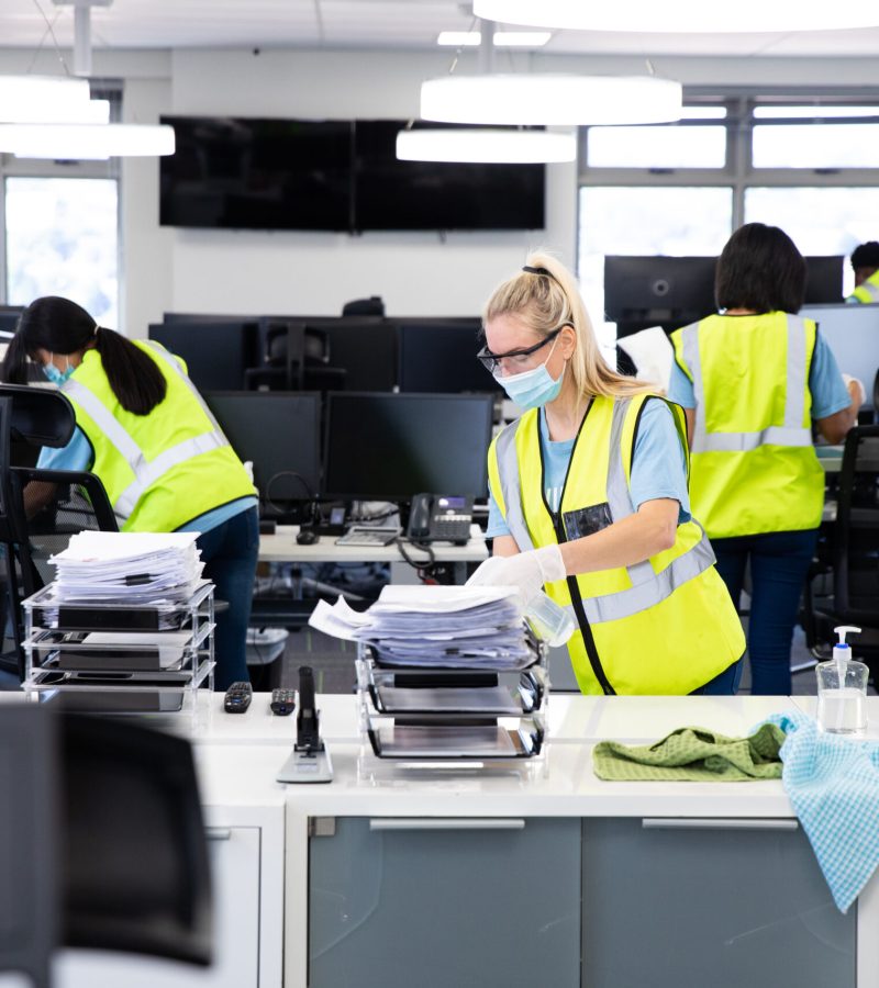 Caucasian woman and colleagues wearing hi vis vests, gloves, safety glasses and face masks sanitizing an office using disinfectant. Hygiene in workplace during Coronavirus Covid 19 pandemic.